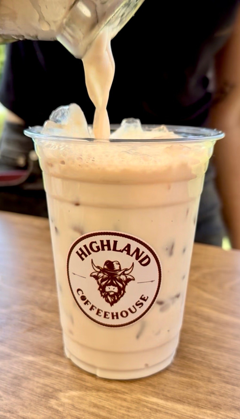 A creamy beverage being poured into a clear cup with Highland Coffeehouse logo, filled with ice on a wooden surface